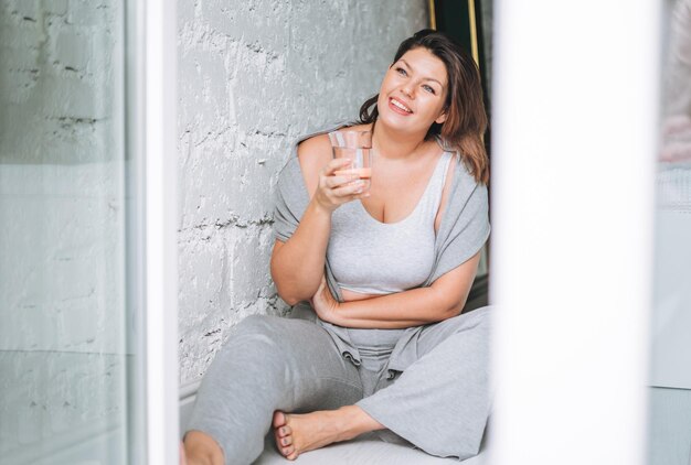 Schöne glückliche junge Frau plus Größenkörper positiv in bequemer Sportkleidung mit Glas Wasser