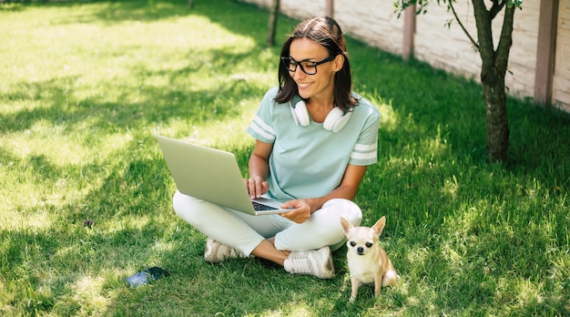 Schöne glückliche junge freiberufliche Frau mit ihrem kleinen niedlichen Hund arbeitet an einem Laptop im Freien.