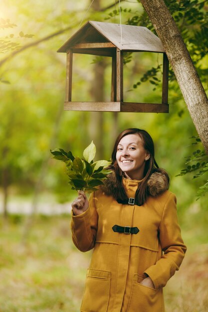 Schöne glückliche kaukasische junge lächelnde braunhaarige Frau im gelben Mantel, in den Jeans, in den Stiefeln im grünen Wald. Mode-Modell mit Herbstlaub, das unter Vogelhäuschen im Frühherbstpark im Freien steht.