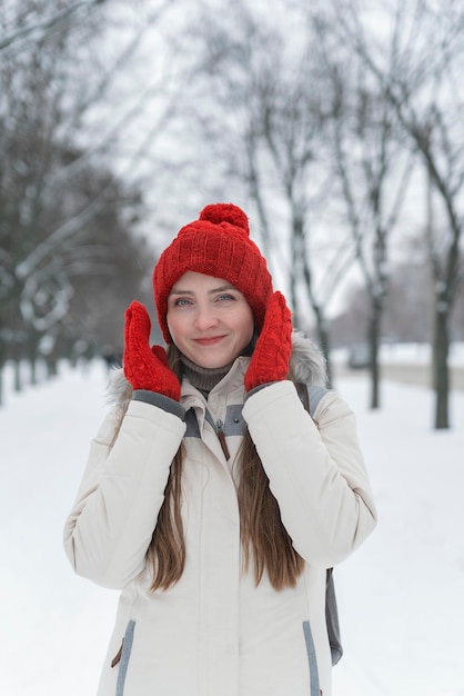 Schöne glückliche lachende junge Frau im roten Hut des Winters und in den Handschuhen nahe dem Gesicht. Winterwaldlandschaftshintergrund.