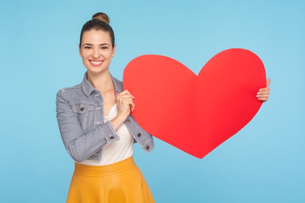 Schöne, glückliche, modisch gekleidete Frau mit liebenswürdigem Lächeln, die ein großes rotes Papierherz hält, das Liebe, Zuneigung, Symbol für Nächstenliebe und Freundlichkeit ausdrückt, Studioaufnahme isoliert auf blauem Hintergrund