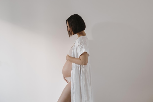 Schöne glückliche schwangere Frau in eleganter weißer Robe, die ihren Bauch berührt Ästhetische elegante Schwangerschaft Mutterschaft Neugeborenes erwartet Konzept
