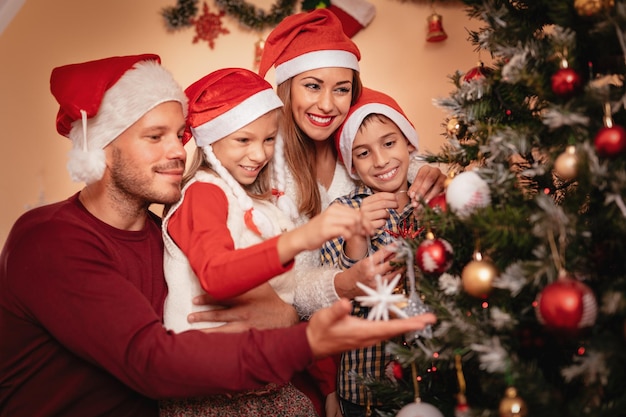 Schöne glückliche Schwester und Bruder schmücken den Weihnachtsbaum mit ihren Eltern zu Hause.