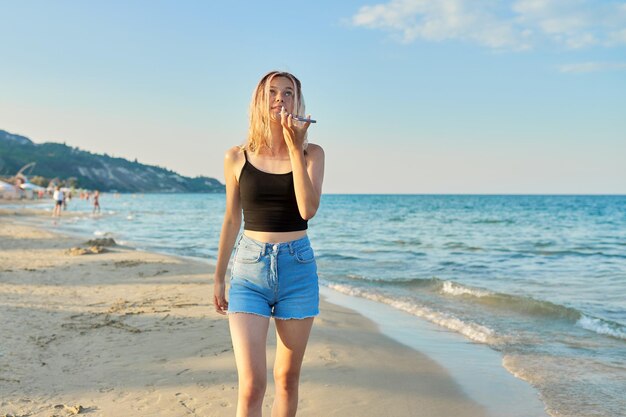 Schöne glückliche Teenagerin mit Smartphone, die bei Sonnenuntergang am Sandstrand entlang spaziert