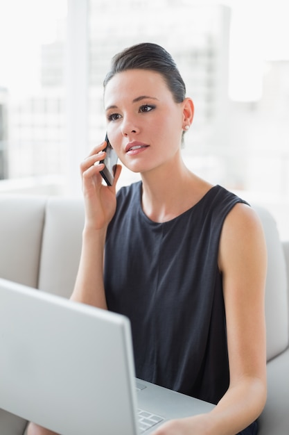 Schöne gut gekleidete Frau, die Laptop und Mobiltelefon auf Sofa verwendet