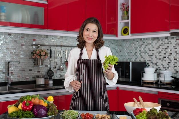 Schöne Hausfrau der asiatischen Frau des mittleren Alters, die Schürze steht, die neue rote Tonküche steht und frisches grünes Gemüse und Messer hält Gemüsesalat hält.