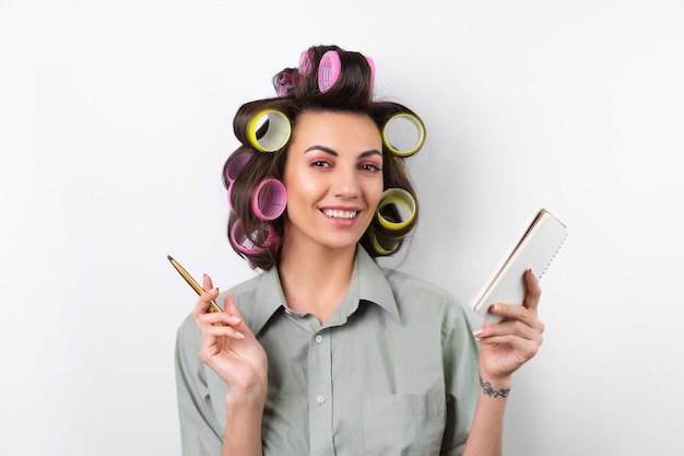 Schöne Hausfrau Junge fröhliche Frau mit hellen Lockenwicklern Make-up ein Notizbuch und einen Schneebesen in ihren Händen auf weißem Hintergrund Nachdenken über das Rezept für das Abendessen