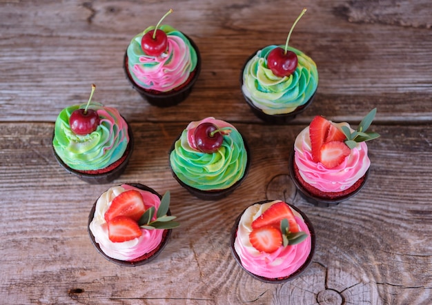 Schöne hausgemachte Cupcakes mit rosa und grüner Käsecreme und Erdbeeren und Kirschen auf einem grauen Holztisch