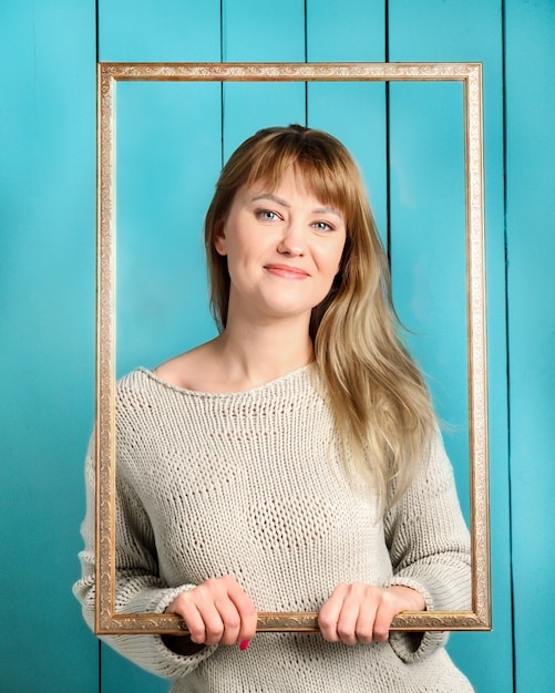 Schöne hübsche blonde junge Frau im gestrickten Pullover mit losen Haaren hält Vintage hölzernen Bilderrahmen und lächelt