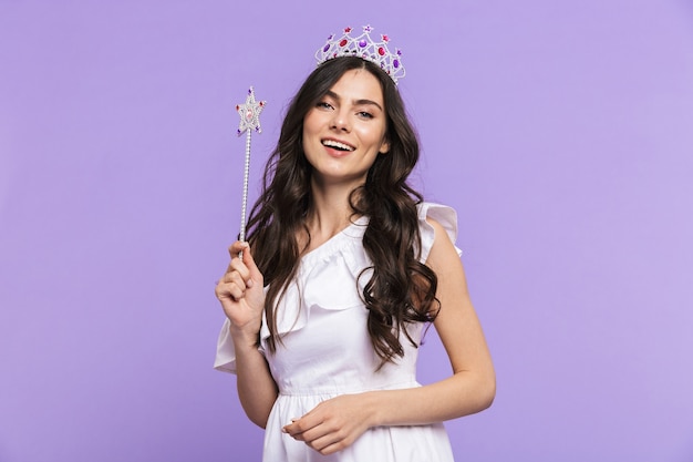 Schöne hübsche junge Frau mit Prinzessinnen-Outfit, die isoliert über der violetten Wand steht und mit magischem Zauberstab posiert