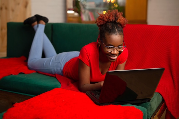 Schöne junge afrikanische frau liegt mit einem laptop auf der couch