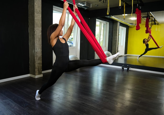 Schöne junge afrikanische Frau mit schlankem Körper, die ihre Beine streckt, während sie fliegende Yogaübungen auf einer roten Hängematte im Fitnessstudio macht