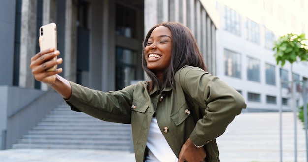 Schöne junge Afroamerikanerfrau, die zur Smartphone-Kamera aufwirft und lächelt, während Selfie-Foto an der Straße nimmt. Ziemlich fröhliche stilvolle Frau, die Selfies Bilder mit Handy macht.