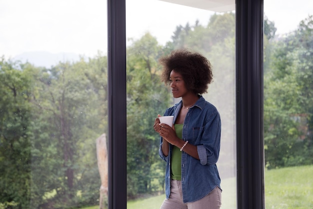 Schöne junge afroamerikanische Frau, die Kaffee trinkt und durch ein Fenster in ihrem Luxushaus schaut
