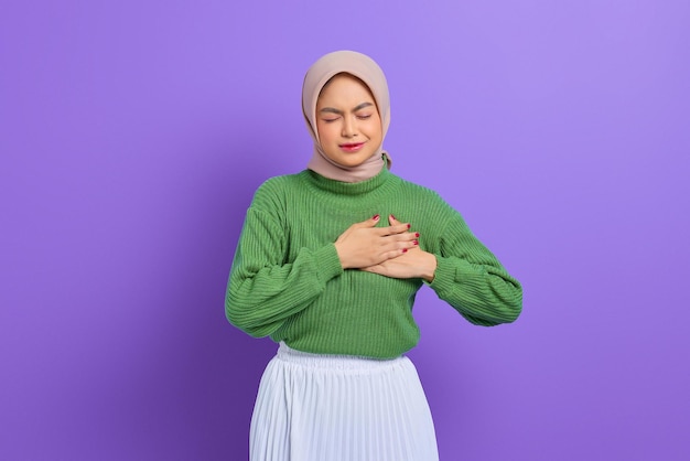 Schöne junge Asiatin im grünen Pullover drückt eine Hand auf die Brust, hat einen Herzinfarkt, der unter unerträglichen Schmerzen leidet, schließt die Augen einzeln auf violettem Hintergrund