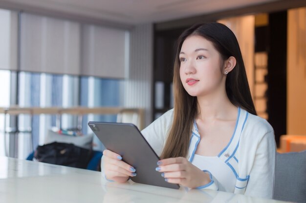 Schöne junge asiatische Berufsfrau mit langen Haaren im weißen Hemd hält Tablette, die glücklich lächelt, während sie auf einem Stuhl im Arbeitsplatzbüro mit verglastem Gebäude als Hintergrund sitzt.