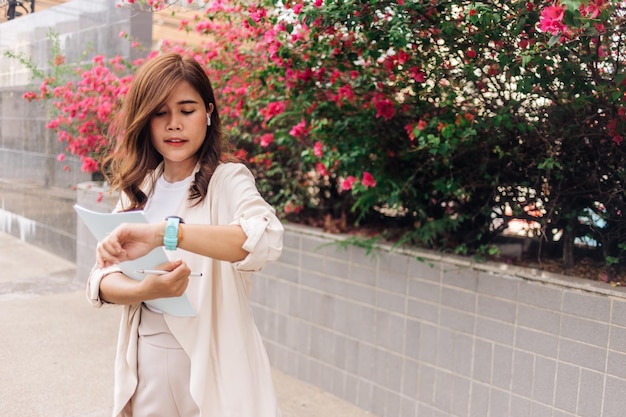 Schöne junge asiatische Frau benutzt eine Smartwatch und liest ein Buch in der Stadt