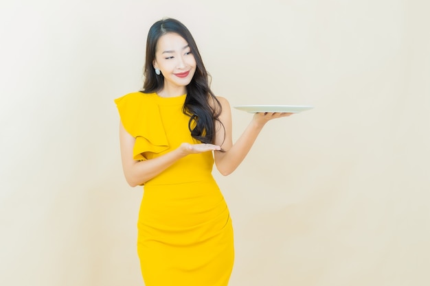 Schöne junge asiatische Frau des Porträts lächelt mit leerem Tellerteller auf beige Wand