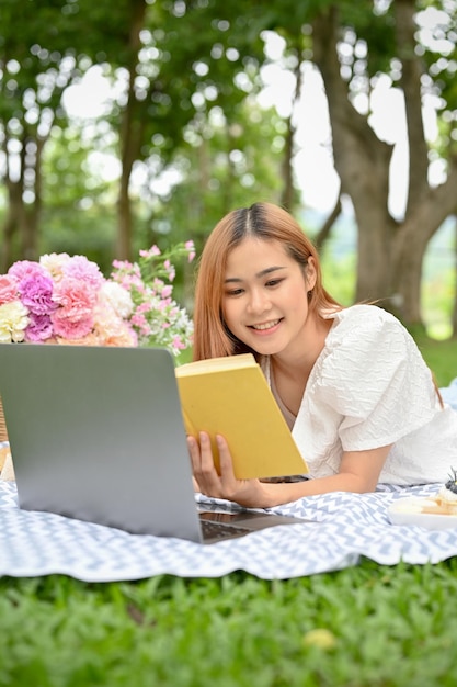 Schöne junge asiatische Frau, die auf der Picknickdecke liegt und ein Buch im Park liest