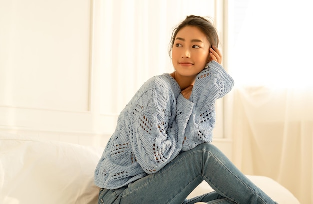 Schöne junge asiatische Frau, die freundlich in gestricktem Pullover lächelt und den sonnigen Abend genießt und wegschaut, während sie auf dem Bett im Schlafzimmer sitzt