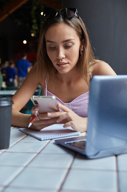 Schöne junge asiatische frau, die mit laptop im café mit smartphone sitzt