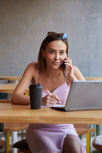 Schöne junge asiatische Frau, die mit Laptop im Café sitzt und mit dem Smartphone anruft