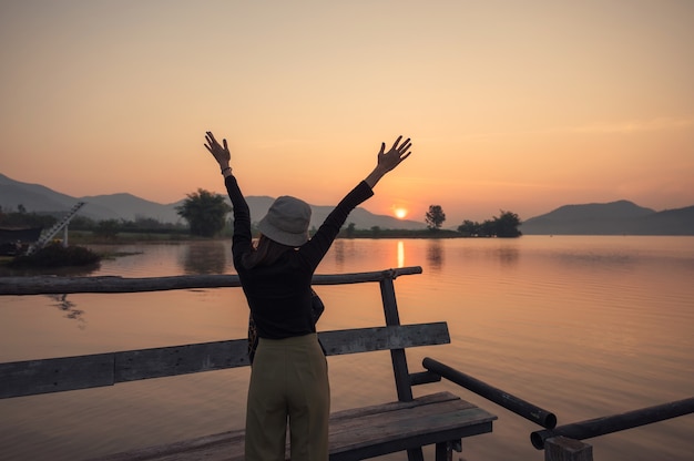 Schöne junge asiatische Frau, die mit Sonnenaufgang auf Bergsee am hölzernen Pier am Morgen genießt