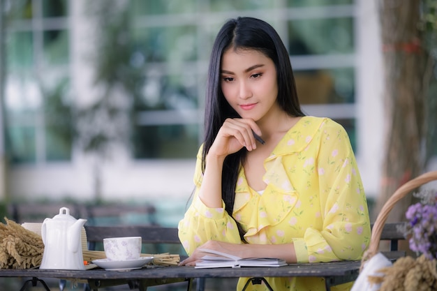 Schöne junge asiatische Frau Hand schreibt auf Notizblock mit einem Stift im Garten und Teetasse mit Blume auf dem Tisch