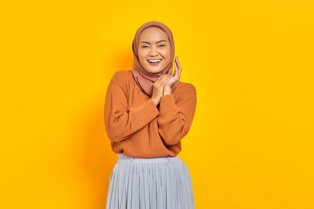 Schöne junge asiatische Frau im braunen Pullover lächelt glücklich und sieht selbstbewusst isoliert auf gelbem Hintergrund aus. Muslimisches Lifestyle-Konzept