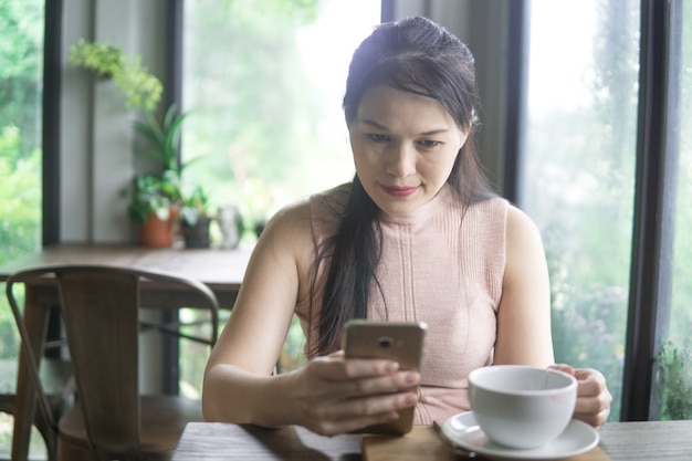 Schöne junge asiatische Frau im Café, unter Verwendung des Handys und des trinkenden Kaffeelächelns.