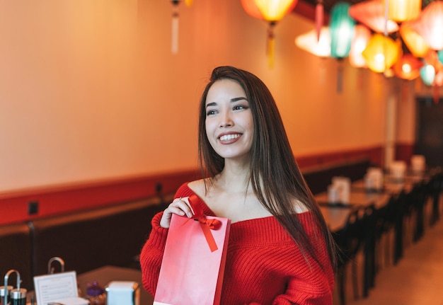 Schöne junge asiatische Frau in roter Kleidung mit Geschenktüte im Restaurant, das das chinesische Neujahr feiert