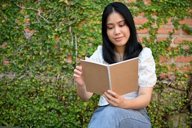 Schöne junge asiatische Frau liest gerne Bücher oder Romane in ihrem schönen Garten oder Hinterhof