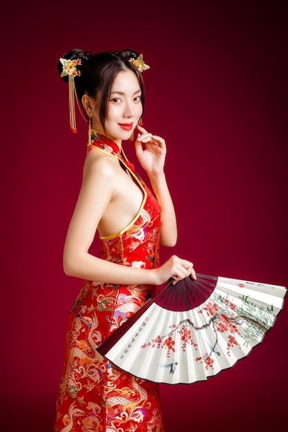 Schöne junge asiatische Frau mit sauberer, frischer Haut, die traditionelles Cheongsam-Qipao-Kleid trägt, das Fan posiert auf rotem Hintergrund hält Porträt des weiblichen Modells im Studio Frohes chinesisches neues Jahr