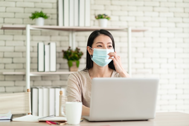 Schöne junge asiatische Frau trägt eine chirurgische Maske, die von zu Hause aus arbeitet und sich mit fröhlichem Ausdruck glücklich lächelt