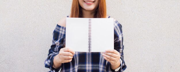 Schöne junge asiatische Frau versteckt sich hinter einem leeren mock up Buch auf Betonwand Hintergrund. Vintage Effekt Stil Bilder. Panorama-Banner.