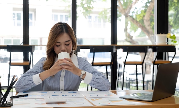 Schöne junge asiatische Geschäftsfrau, die eine Kaffeetasse und einen Laptop hält, die im Büro arbeiten