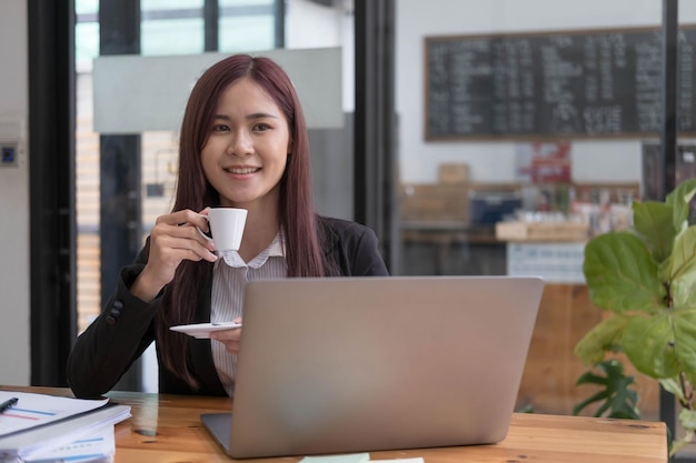 Schöne junge asiatische Geschäftsfrau, die eine Kaffeetasse und einen Laptop hält, die im Büro arbeiten