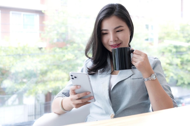 Schöne junge asiatische Geschäftsfrau, die im Büro lächelt und eine Kaffeetasse und ein Smartphone hält