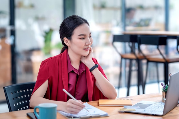 Schöne junge asiatische Geschäftsfrau glücklich im Büro mit der Schreibarbeit, die einen Stift hält.