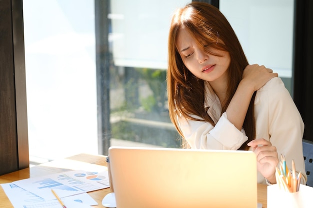 Schöne junge asiatische Geschäftsfrau mit Bürosyndrom hat ein Problem mit einem Teil des Körpers, Arm, Schulter, Kopf, Nacken, Rücken, gesundes Problem, nachdem sie lange am Laptop gearbeitet hat.