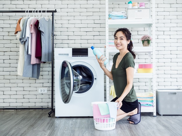Schöne junge asiatische Hausfrau der Frau, die flüssiges Waschmittel, blaue Flasche mit dem Lächeln und Betrachten der Kamera nahe der Waschmaschine im Waschraum sitzt und hält.