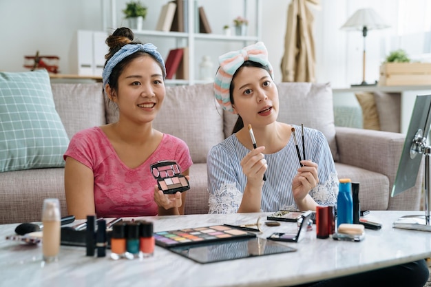 Schöne junge asiatische Koreanerinnen mit Stirnbändern, die zu Hause Schönheitsprodukte im Videoblog überprüfen. freundinnen vlogger lehren zusammen make-up, das kosmetikprodukt-lidschatten zur kamera drinnen zeigt.