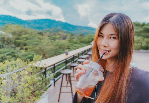 Schöne junge asiatische lächelnde Frau ist im Café mit Wald- und Bergnaturhintergrund, während sie eisgekühlten Americano-Kaffee trinkt