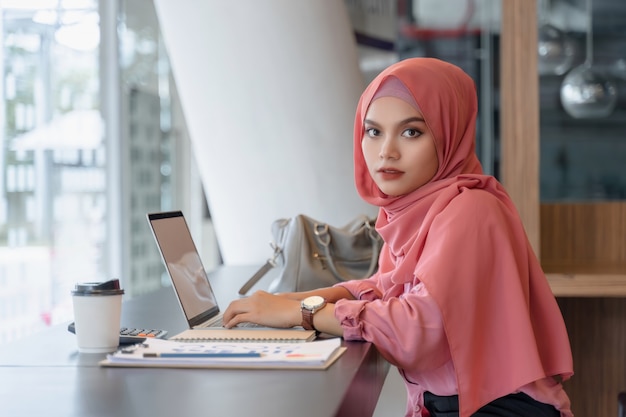 Schöne junge asiatische moslemische Geschäftsfrau im rosa hijab und in der Freizeitkleidung, die mit Laptop arbeitet