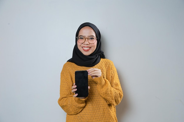 Schöne junge asiatische Muslimin mit orangefarbenem Pullover und Brille, die ihr Telefon in die Kamera zeigt