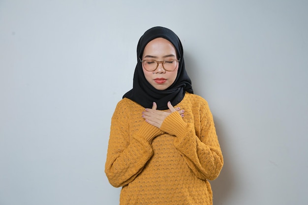 Schöne junge asiatische muslimische Frau mit orangefarbenem Pullover und Brille, die die Brust hält