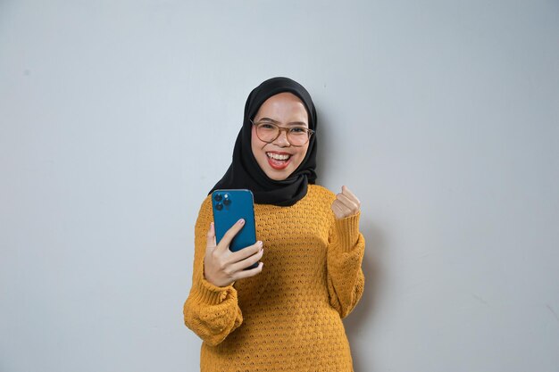 Schöne junge asiatische muslimische Frau trägt orangefarbenen Pullover und Brille, während sie ihr Mobiltelefon benutzt