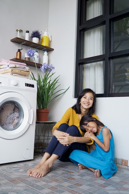 Schöne junge asiatische Mutter und Tochter warten auf die Wäschewaschmaschine, um das Spinnen zu beenden