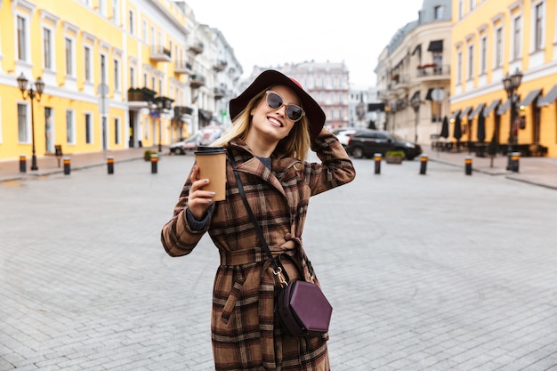 Schöne junge blonde Frau, die einen Mantel trägt, der draußen geht und Kaffeetasse zum Mitnehmen hält