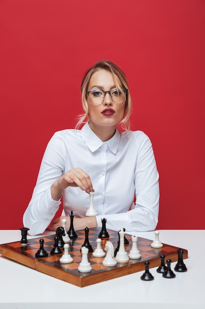 Schöne junge blonde Frau, die weißes Hemd sitzt, das am Tisch sitzt und Schach lokalisiert über rotem Hintergrund spielt